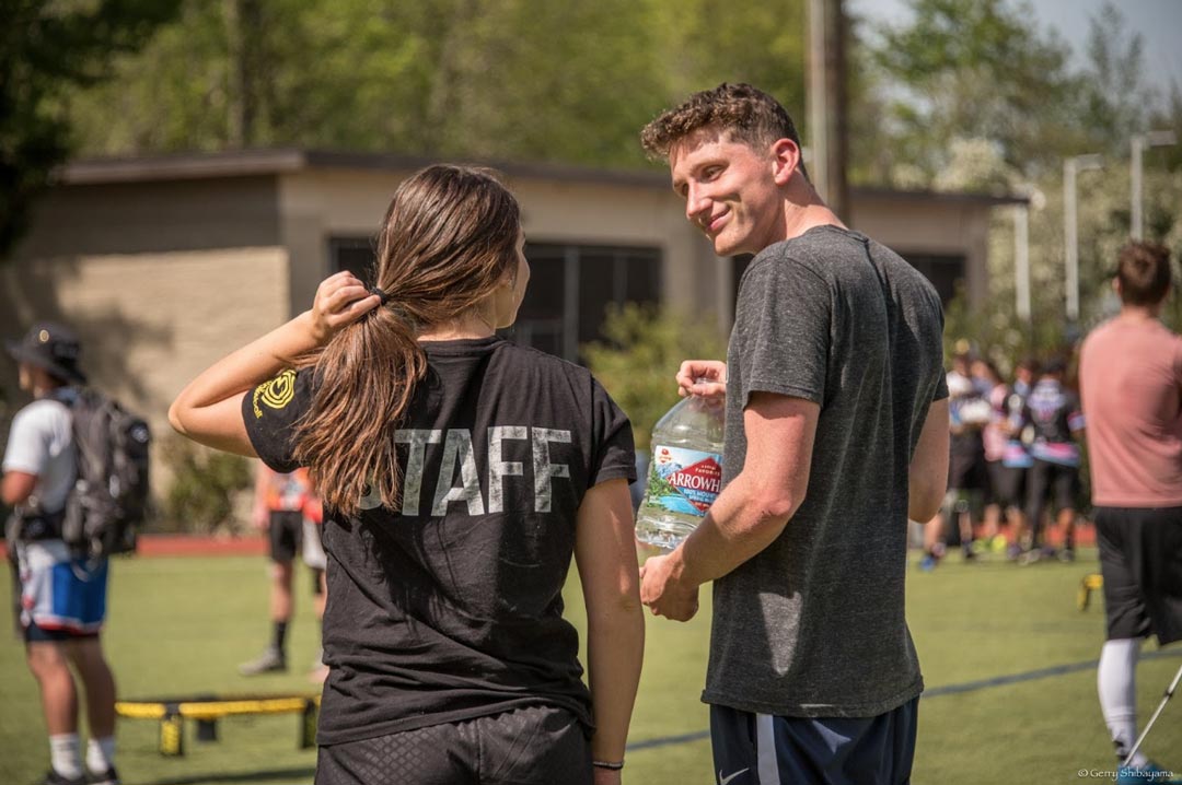 Un homme sourit à une femme portant un t-shirt noir « STAFF » alors qu'ils se trouvent sur un terrain de Spikeball herbeux, d'autres joueurs étant visibles en arrière-plan.