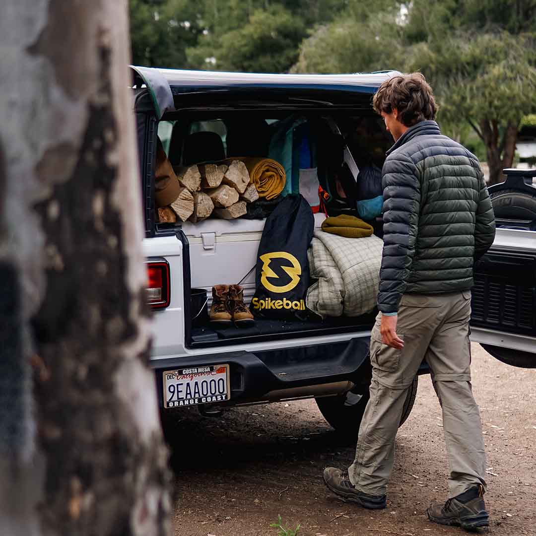 Un homme charge le hayon de son VUS avec du matériel de camping, notamment un sac de Spikeball, du bois de chauffage, une couverture et des bottes, sur un terrain de camping boisé.