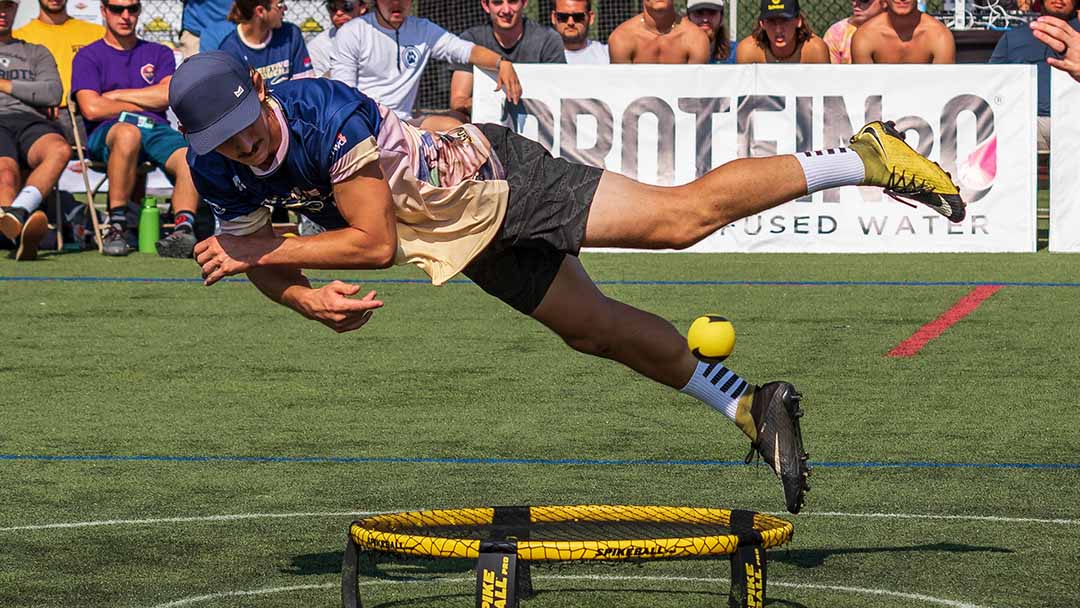 Un joueur plonge pour frapper une Spikeball jaune sur un terrain de roundnet lors d'un tournoi extérieur, avec des spectateurs en arrière-plan.