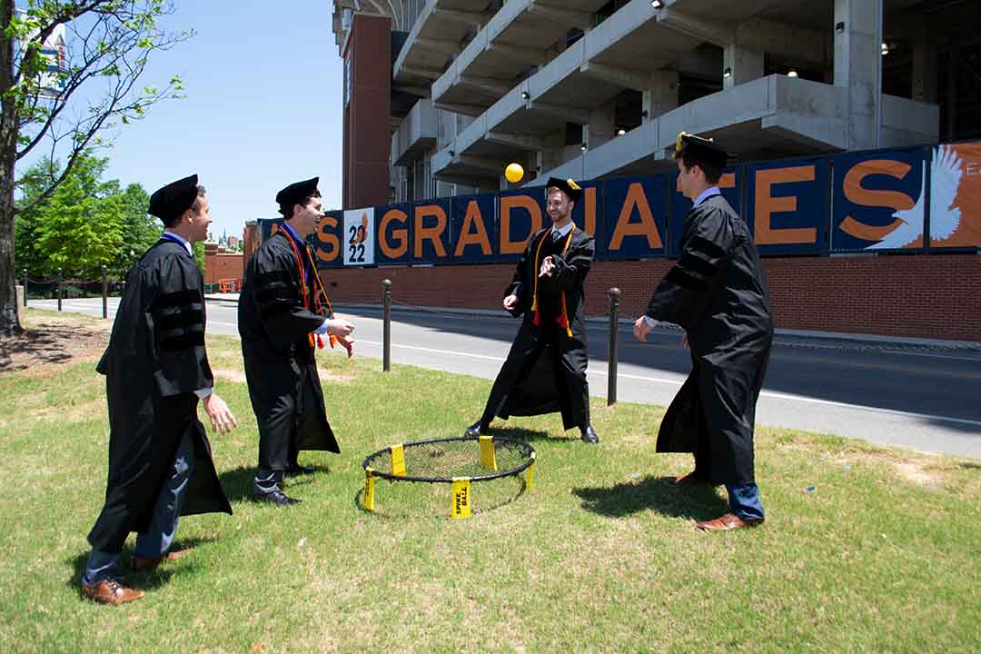 Quatre diplômés en toque et en robe de cérémonie jouent au Spikeball sur l'herbe à côté d'un stade orné d'une grande bannière « DIPLÔMÉS ».