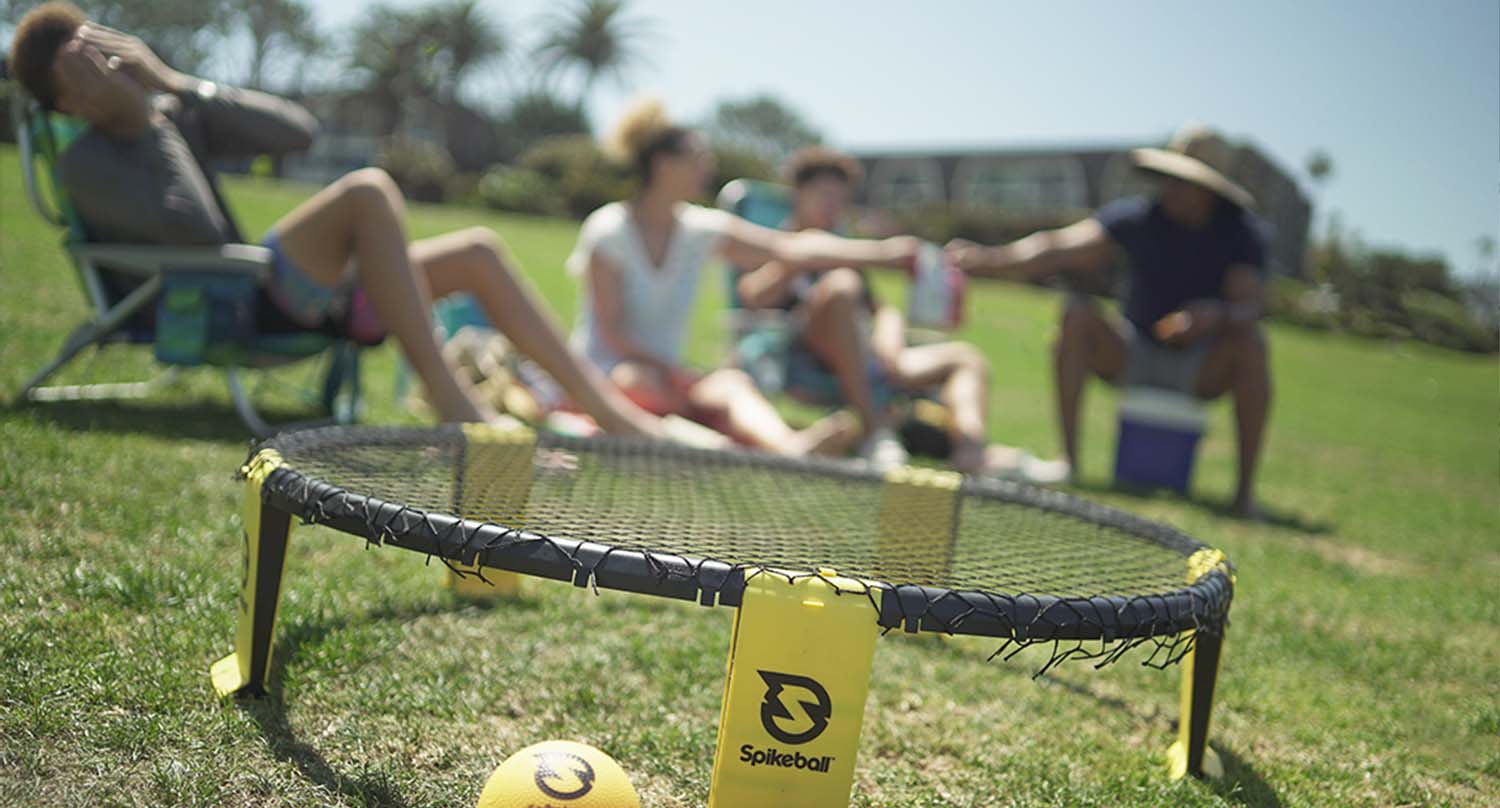 Filet de spikeball sur l'herbe, pieds jaunes et balle au premier plan, quatre personnes se détendant sur des chaises de jardin dans un parc ensoleillé