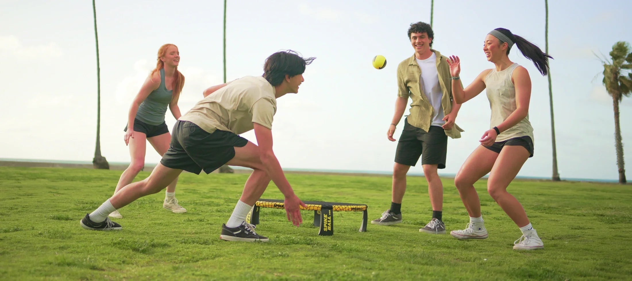 Quatre amis jouent au Spikeball sur une pelouse de plage. L'un d'eux s'accroupit pour renvoyer une balle jaune dans le filet.
