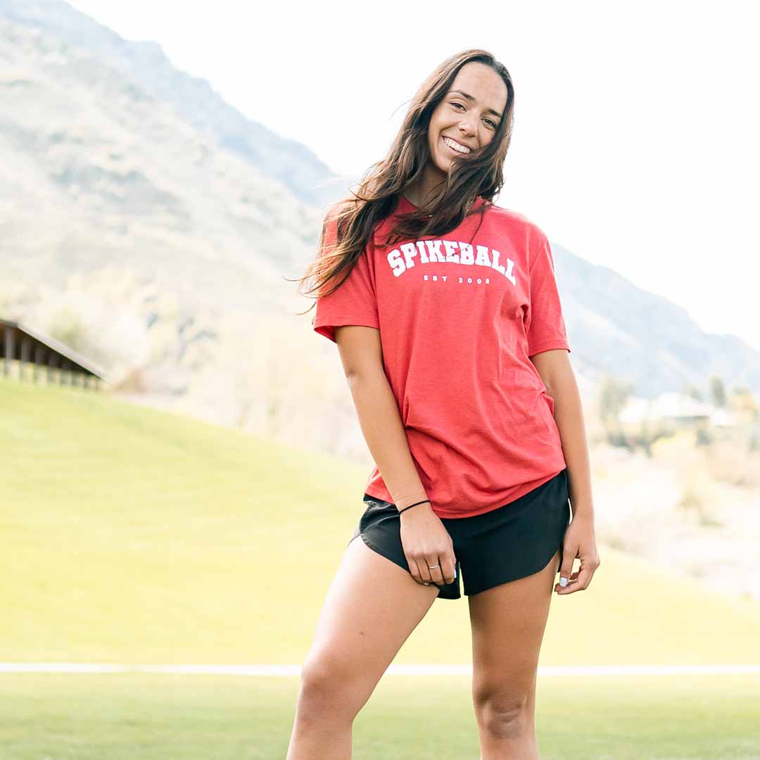 Front view of red Spikeball tee with white SPIKEBALL EST 2008 chest logo worn by smiling woman on grassy field