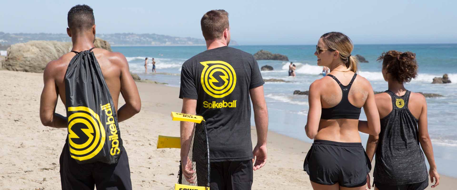 Vier Personen, die mit Spikeball-Ausrüstung am Strand spazieren gehen und schwarze Kleidung mit dem Spikeball-Logo tragen