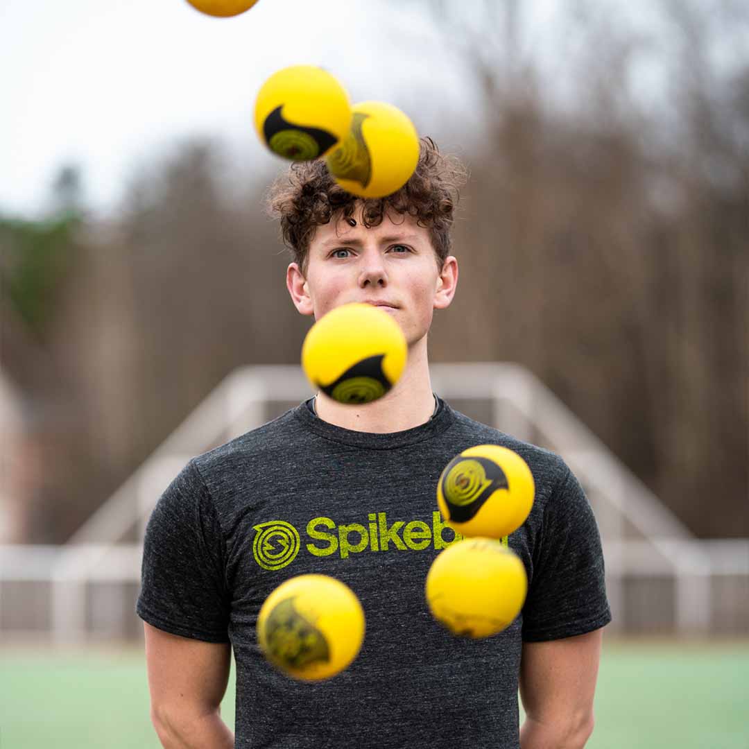 Un jeune homme jongle avec plusieurs balles de Spikeball jaunes sur un terrain de sport extérieur, portant un t-shirt foncé de Spikeball