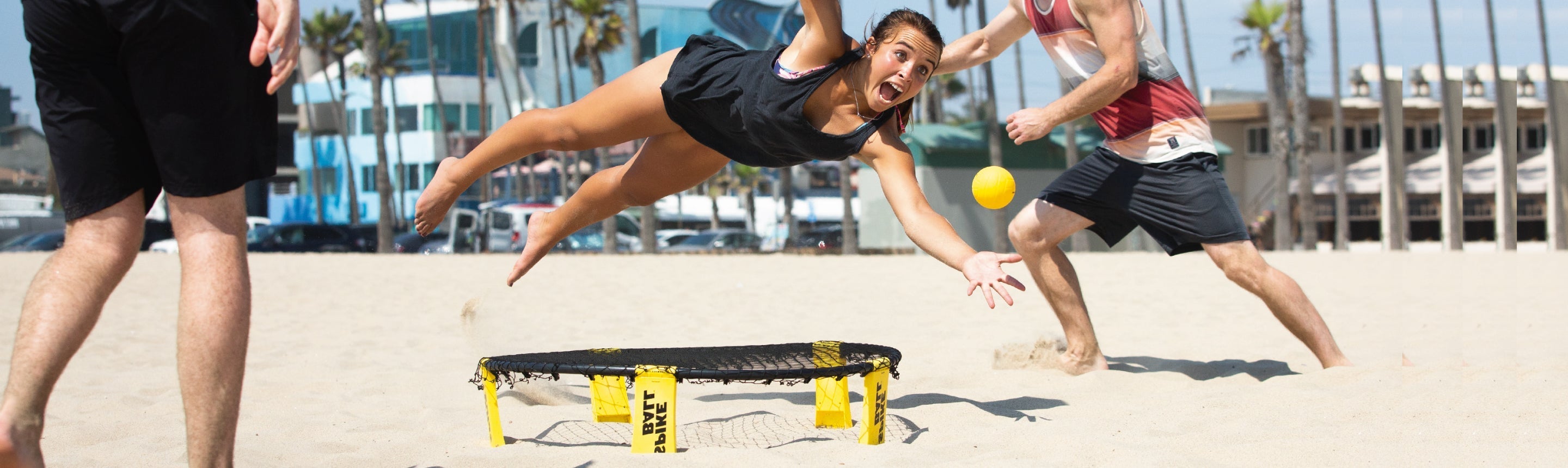 Tres jugadores lanzándose a por una pelota amarilla de Spikeball durante un partido en una playa soleada, con una red de Spikeball en primer plano
