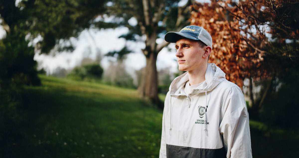 Young man wearing blue and gray Spikeball cap and white windbreaker standing in a grassy park with trees