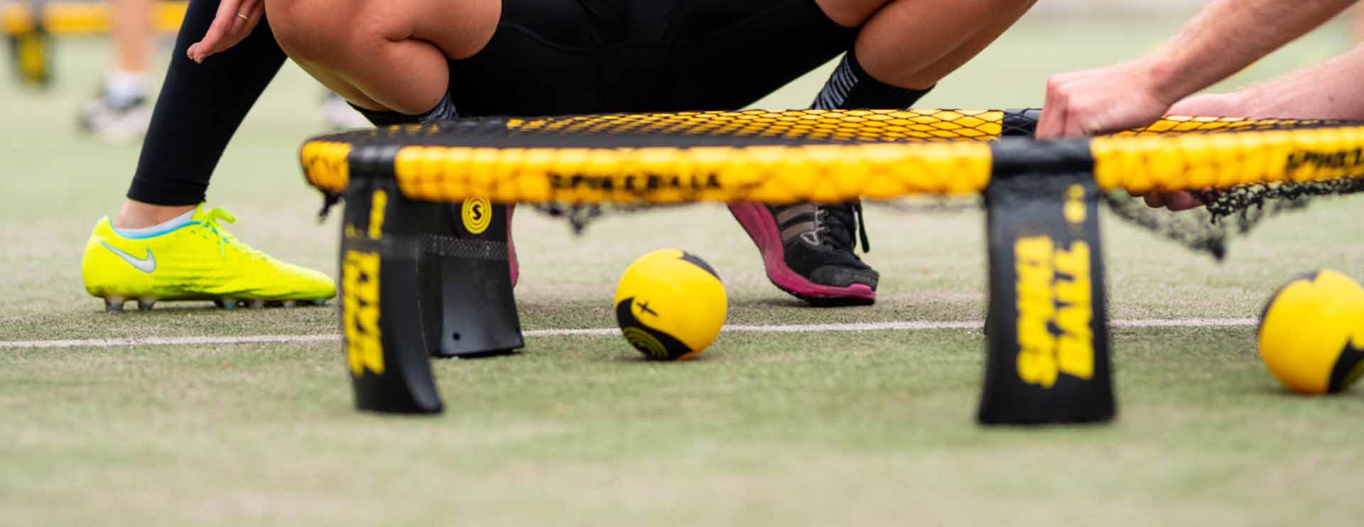 Players setting up a yellow Spikeball net on a grass field with yellow balls and players’ legs visible