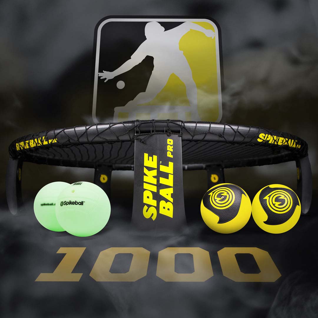 Vista frontale del set Spikeball Pro che mostra il cerchio e la rete neri con due palline da gioco gialle e nere e due palline da allenamento verde fosforescente su uno sfondo scuro fumé con la scritta “1000” in oro