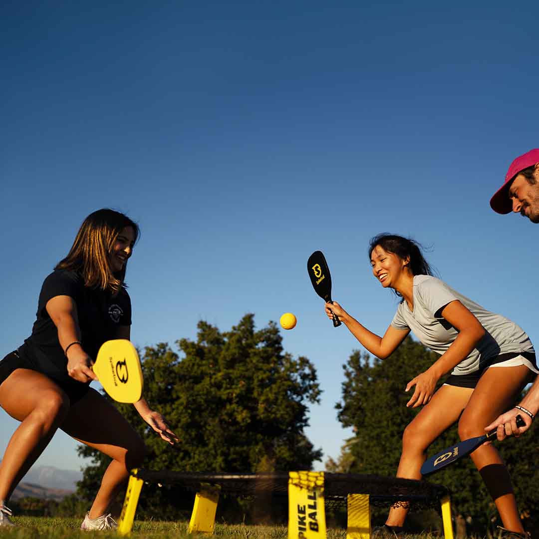 Two women and a man playing Spikeball with paddles on a grassy field under a clear blue sky. The players are actively hitting the yellow ball, and a Spikeball net is set up on the ground.