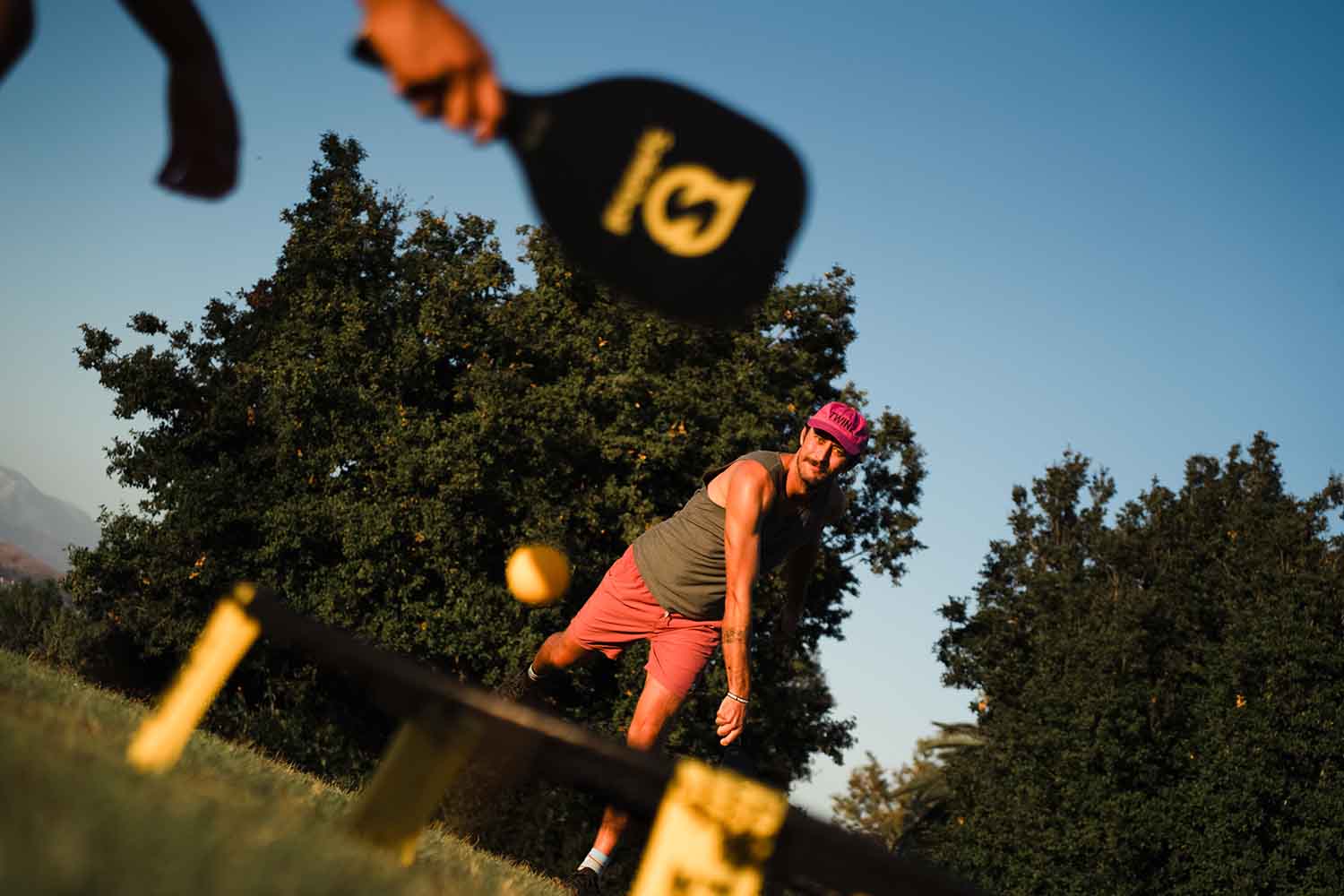 Un joueur utilise une raquette SpikePaddle pour frapper une balle à pointes jaune vers un filet bas sur un terrain herbeux bordé d'arbres et sous un ciel bleu.